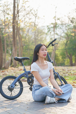 Young asian women wearing earphone and listening audio music on smartphone with happiness while sitting on the street to relaxation with journey travel lifestyle after cycling bicycle in the park.の写真素材