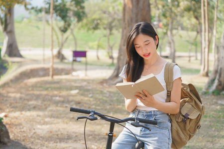 Young asian women with backpack sitting on bicycle and reading a book with happiness to resting after cycling bicycle in the nature park while relaxation with journey travel for healthy lifestyle.の写真素材