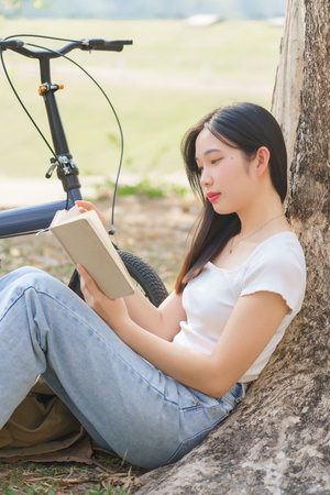 Young asian women sitting on grass to leaning the tree and reading a book with recreation happiness while relaxation with journey travel trip lifestyle after cycle bicycle in the nature park.の写真素材