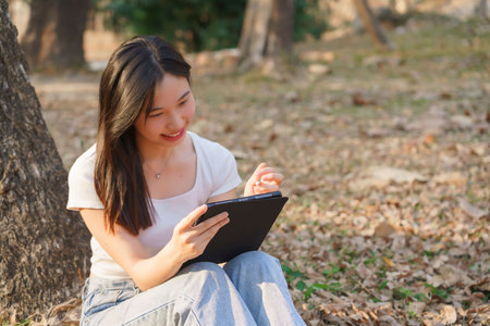 Young asian women sitting on the grass under tree in the nature park and using digital tablet to writing business information while working outside and relaxation with journey travel lifestyle.の写真素材