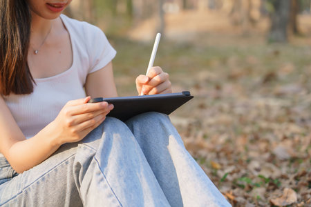 Young asian women writing business data and journey diary on digital tablet while sitting on the grass under tree in the nature park to working outside and relaxation with journey travel lifestyle.の写真素材