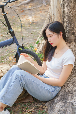 Young asian women sitting on grass to leaning the tree and reading a book with recreation happiness while relaxation with journey travel trip lifestyle after cycle bicycle in the nature park.の写真素材