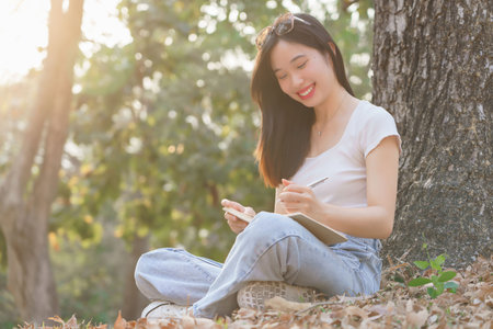 Young asian women sitting on grass under the tree to writing business data and journey dairy in notebook while enjoying and happiness to relaxation with journey travel lifestyle in the nature park.の写真素材