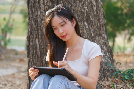Young asian women writing business data and journey diary on digital tablet while sitting on the grass under tree in the nature park to working outside and relaxation with journey travel lifestyle.の写真素材