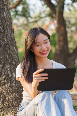 Young asian women sitting on the grass under tree in the nature park and using digital tablet to writing business information while working outside and relaxation with journey travel lifestyle.の写真素材