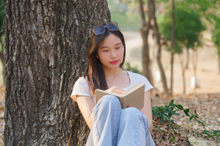 Young asian women sitting on grass to leaning the tree and reading a literature book with recreation happiness while enjoying to relaxation with journey travel trip lifestyle in the nature park.の写真素材