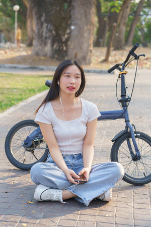 Young asian women wearing earphone to listening music on smartphone and enjoying with nature view while sitting on the street to relaxation with journey travel lifestyle after cycling bicycle in park.の写真素材