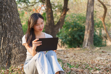 Young asian women writing business data and journey diary on digital tablet while sitting on the grass under tree in the nature park to working outside and relaxation with journey travel lifestyle.の写真素材