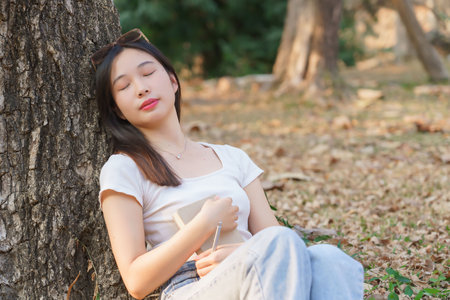 Young asian women sitting on grass and to leaning the tree and sleeping with dreaming to resting after reading a literature book while relaxation with journey travel lifestyle in the nature park.の写真素材