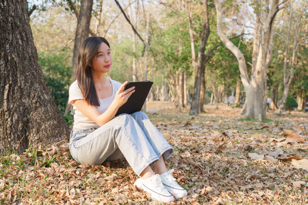Young asian women sitting on the grass under tree in the nature park and using digital tablet to writing business information while working outside and relaxation with journey travel lifestyle.の写真素材