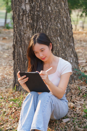 Young asian women sitting on the grass under tree in the nature park and using digital tablet to writing business information while working outside and relaxation with journey travel lifestyle.の写真素材