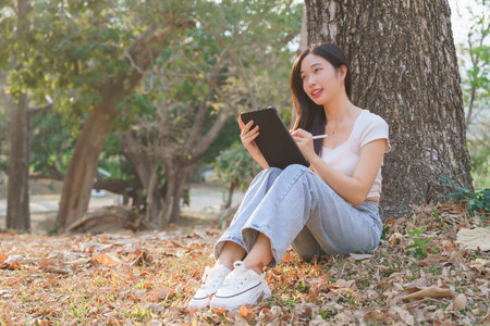 Young asian women sitting on the grass under tree in the nature park and using digital tablet to writing business information while working outside and relaxation with journey travel lifestyle.の写真素材