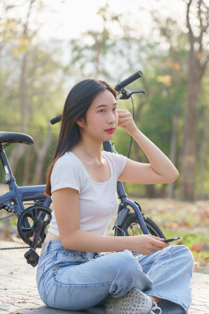 Young asian women wearing earphone and listening audio music on smartphone with happiness while sitting on the street to relaxation with journey travel lifestyle after cycling bicycle in the park.の写真素材