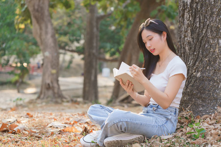 Young asian women sitting on grass under the tree to writing business data and journey dairy in notebook while enjoying and happiness to relaxation with journey travel lifestyle in the nature park.の写真素材