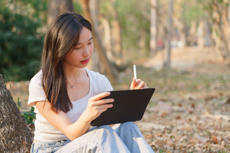 Young asian women writing business data and journey diary on digital tablet while sitting on the grass under tree in the nature park to working outside and relaxation with journey travel lifestyle.の写真素材
