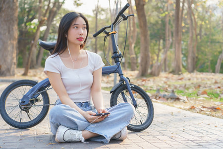Young asian women wearing earphone and listening audio music on smartphone with happiness while sitting on the street to relaxation with journey travel lifestyle after cycling bicycle in the park.の写真素材