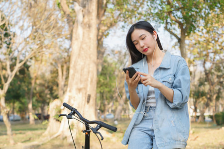 Young asian women wearing earphone and using smartphone to listening audio music and chatting while sitting on bicycle to relaxation with journey travel lifestyle after cycling bicycle in the park.の写真素材