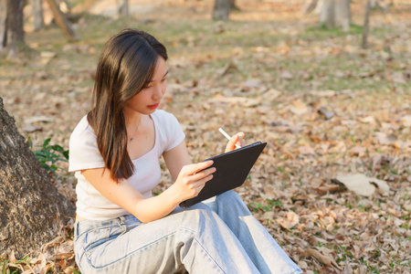 Young asian women writing business data and journey diary on digital tablet while sitting on the grass under tree in the nature park to working outside and relaxation with journey travel lifestyle.の写真素材