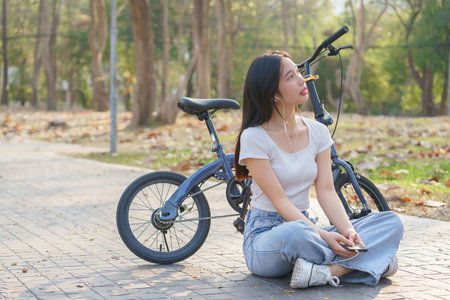 Young asian women wearing earphone and listening audio music on smartphone with happiness while sitting on the street to relaxation with journey travel lifestyle after cycling bicycle in the park.の写真素材