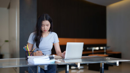 Asian teenage student woman reading a book to taking notes for assignment homework while watching lecture video online lesson on laptop to studying and learning about education knowledge in cafe.の写真素材