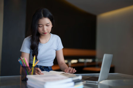 Asian teenage student woman reading a book and writing notes to doing homework while watching lecture video online lesson on digital laptop to studying and learning about education knowledge in cafe.の写真素材