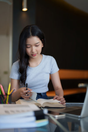 Asian teenage student woman reading a book and writing notes to doing homework while watching lecture video online lesson on digital laptop to studying and learning about education knowledge in cafe.の写真素材