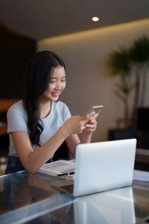 Asian teenage student woman using smartphone to chatting with friends and doing assignment homework after watching lecture video online lesson on laptop and studying about education knowledge in cafe.の写真素材