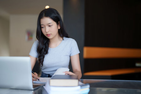 Asian teenage student woman reading a book to taking notes for assignment homework while watching lecture video online lesson on laptop to studying and learning about education knowledge in cafe.の写真素材