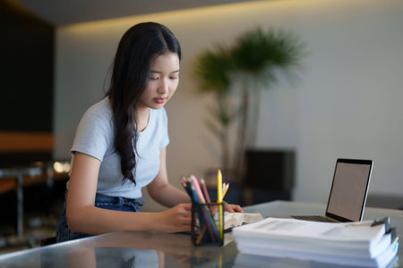 Asian teenage student woman reading a book to writing information from book and doing assignment homework after watching video online lesson on laptop and studying about education knowledge in cafe.の写真素材