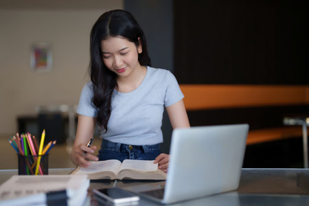 Asian teenage student woman reading a book and writing notes to doing homework while watching lecture video online lesson on digital laptop to studying and learning about education knowledge in cafe.の写真素材