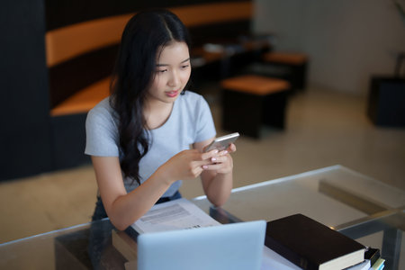 Asian teenage student woman researching information on smartphone and doing assignment homework after watching lecture video online lesson on laptop and studying about education knowledge in cafe.の写真素材