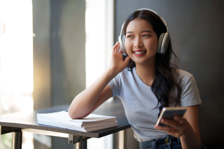 Asian teenage student woman wearing headphone to listening music and surfing social media on smartphone to relaxation with happiness after studying education knowledge for preparing test exam in cafe.の写真素材