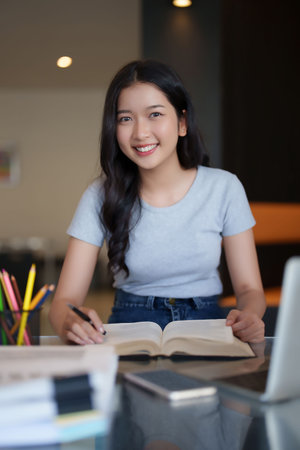 Asian teenage student woman reading a book to writing information from book and doing assignment homework after watching video online lesson on laptop and studying about education knowledge in cafe.の写真素材