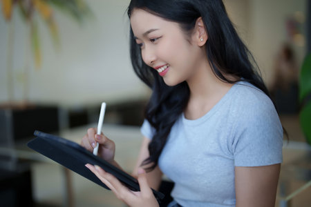 Asian teenage student woman watching video online lesson and writing notes on digital tablet while studying about education knowledge and doing assignment homework for preparing test exam in cafe.の写真素材