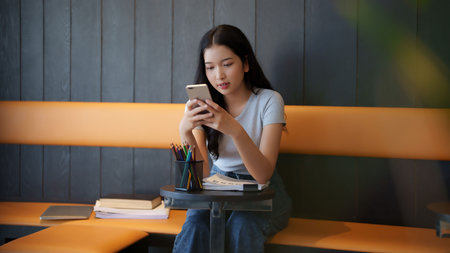 Asian teenage student woman using smartphone to chatting with friends and doing assignment homework after studying and reading information in paperwork to learning about education knowledge in cafe.の写真素材