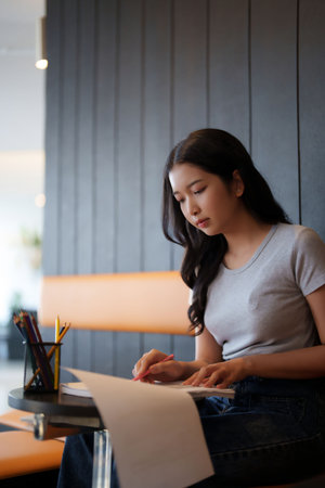 Asian teenage student woman reading information in paperwork to taking notes and doing assignment homework for preparing test exam while studying and learning about education knowledge in cafe.の写真素材