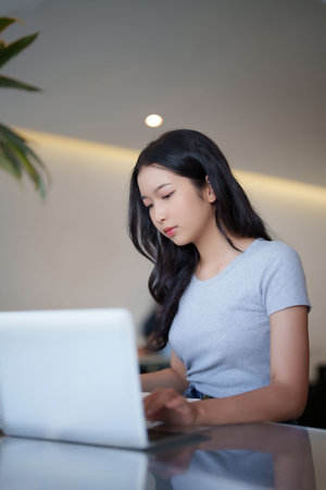 Asian teenage student woman typing and researching data on laptop for doing assignment homework after reading a book and watching lecture video online lesson to studying education knowledge in cafe.の写真素材