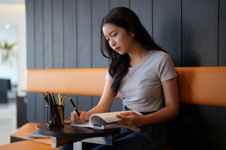 Asian teenage student woman reading a book and writing information from book to doing assignment homework for preparing test exam while studying and learning about education knowledge in cafe.の写真素材