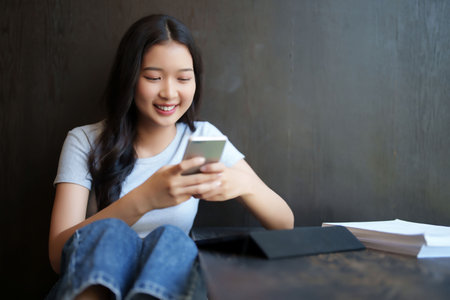Asian teenage student woman using smartphone to surfing social media and chatting with friends with happiness after studying and reading paperwork to learning about education knowledge in cafe.の写真素材