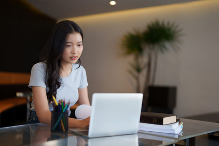 Asian teenage student woman reading a book and typing to researching data on laptop for doing...