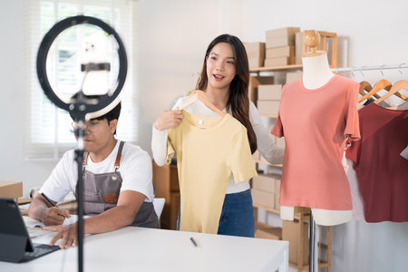 A woman presents a shirt on a mannequin during a livestream while a man works on a laptop in a busy ecommerce workspace.の写真素材