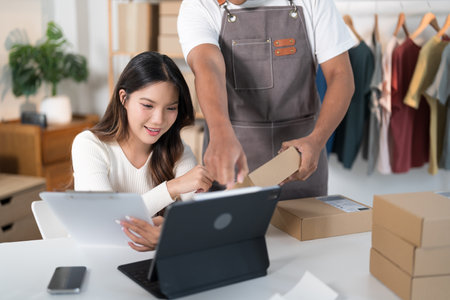 A man packs a box while a woman checks her phone in a workspace with clothing racks and shipping supplies.の写真素材