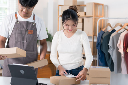 A woman prepares a package for shipping, while another person scans a label in a workspace filled with boxes and clothing racks.の写真素材