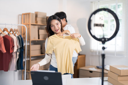 A young woman livestreams showcasing a shirt to her online audience using a smartphone and ring light, with packaging materials and clothing racks in the background.の写真素材