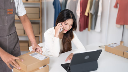 A woman talks on the phone while working on a laptop in a workspace surrounded by boxes and clothing racks.の写真素材