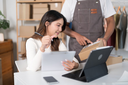 A man packs a box while a woman checks her phone in a workspace with clothing racks and shipping supplies.の写真素材