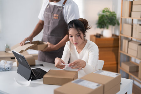 A woman prepares a package for shipping, while another person scans a label in a workspace filled with boxes and clothing racks.の写真素材