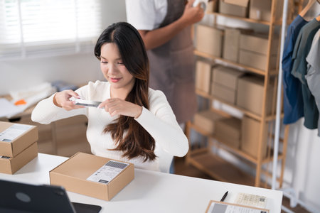 A woman photographs a product on a desk in a workspace filled with shipping boxes and clothing racks.の写真素材