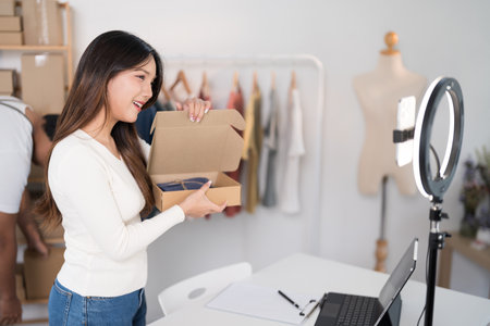 A woman enthusiastically livestreams while holding a box, showcasing her products with a ring light and laptop setup in a workspace filled with racks and packaging.の写真素材