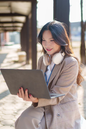 Asian businesswoman working on a laptop while sitting in an outdoor urban setting with headphones around her neckの写真素材
